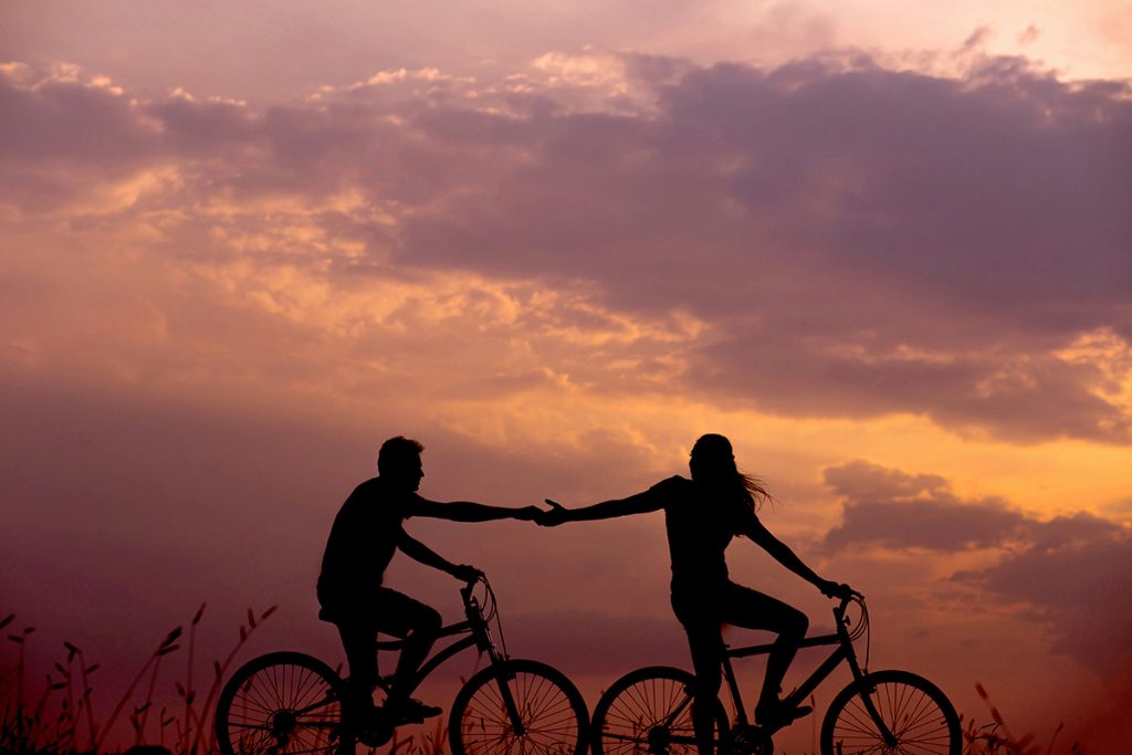 silhouette of two cyclists holding out hands to each other against sunset
